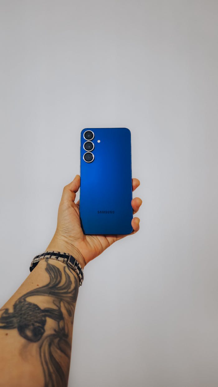 A tattooed hand holding a sleek blue smartphone against a neutral background.